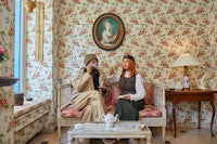 two women sitting on a couch in a room with floral wallpaper